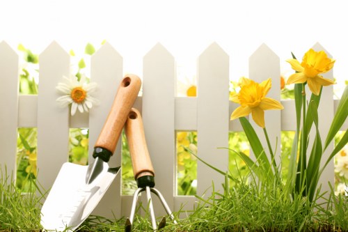 A garden scene featuring a white picket fence in the background with bright yellow daffodils and a white daisy growing alongside lush green grass in the foreground. Various small gardening tools, including a trowel, pruning shears, and a hand rake with wooden handles, are partially embedded in the soil at the edge of the lawn, indicating active gardening or maintenance. The environment appears well-maintained, with a natural and vibrant outdoor space typical of a domestic garden in Regents Park, London. The lighting suggests a clear day with soft sunlight illuminating the plants and garden elements, highlighting the fresh growth and tidy arrangement. This scene reflects a typical landscaped front or backyard designed for both aesthetic appeal and outdoor leisure, associated with professional gardening services in the Regents Park area.
