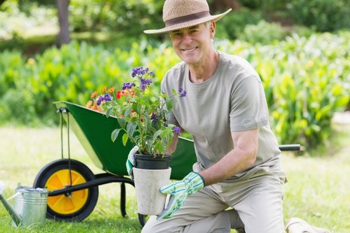 A middle-aged man dressed in light grey gardening attire, including a wide-brimmed hat and gardening gloves, is kneeling on a neatly maintained lawn in an outdoor garden space. He is holding a small potted flowering plant with purple, red, and yellow blooms, which he has just cultivated or prepared for planting. Behind him, there is a green wheelbarrow with a yellow wheel, positioned on the grass, likely used for transporting garden tools and plants. The surrounding environment features a lush, green backdrop of trees and shrubs, with a well-defined flower bed border along the edge of the lawn. The scene appears to be set on a bright, sunny day, with natural light illuminating the vivid colors of the plants and foliage. This outdoor space demonstrates a cared-for, landscaped garden area suitable for gardening services focused on lawn and plant maintenance in Regents Park, London, where natural tones and structured plant arrangements contribute to a serene and inviting environment.