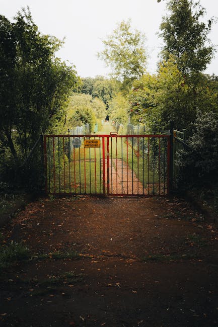 A rust-colored metal gate blocks a narrow, well-trodden dirt pathway leading into a lush green garden area. Surrounding the gate are dense bushes and tall trees with vibrant green foliage, creating a
