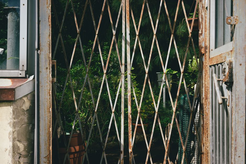 A rust-colored metal gate blocks a narrow, well-trodden dirt pathway leading into a lush green garden area. Surrounding the gate are dense bushes and tall trees with vibrant green foliage, creating a